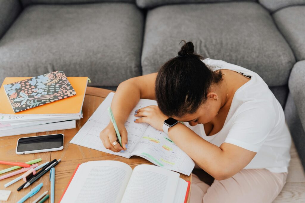 A woman with dark hair leaning her head down on her arms on a wooden table covered in books and papers.