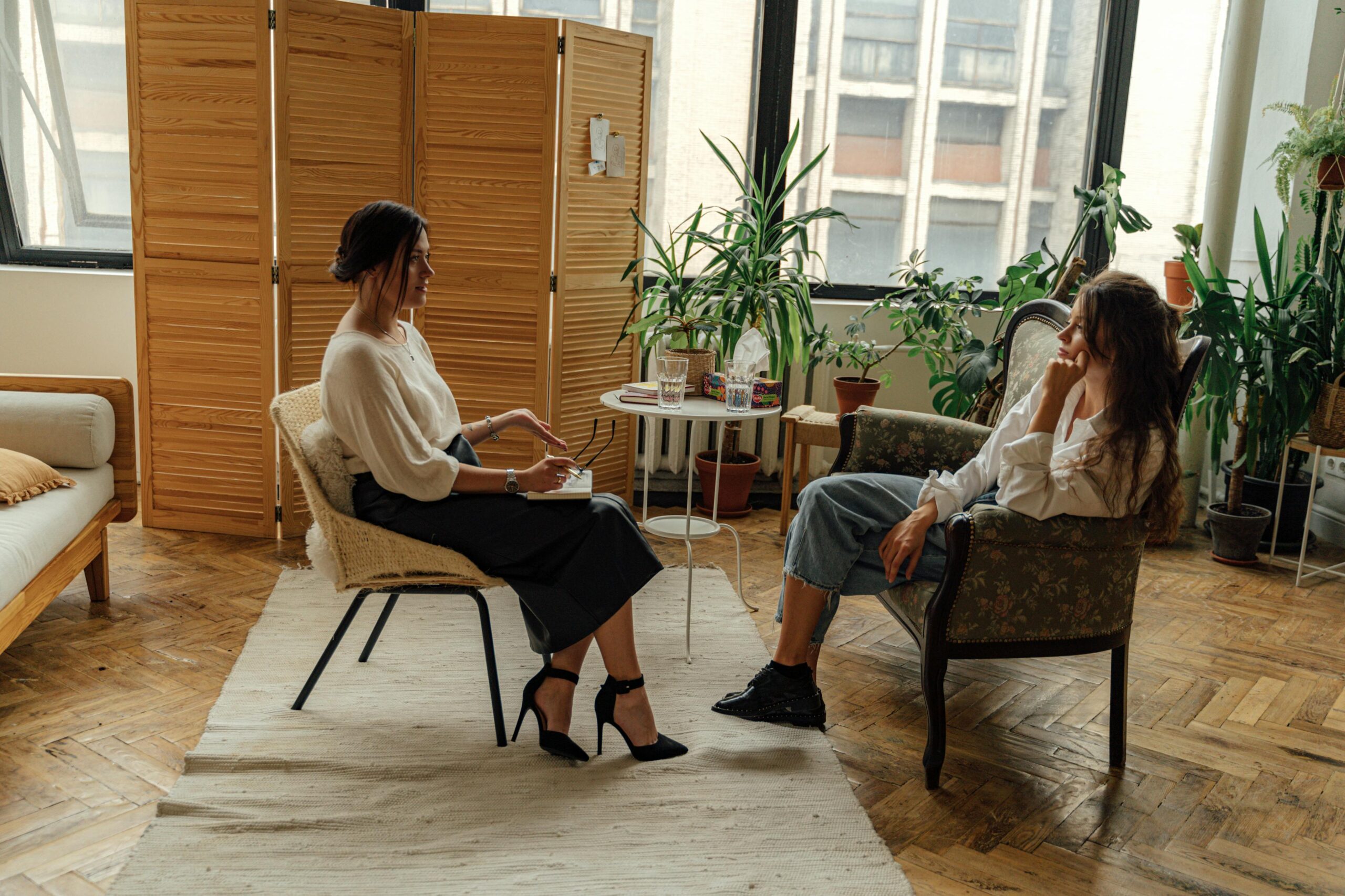 Two women sitting in comfortable chairs in a light-filled therapy office, engaged in a collaborative session.