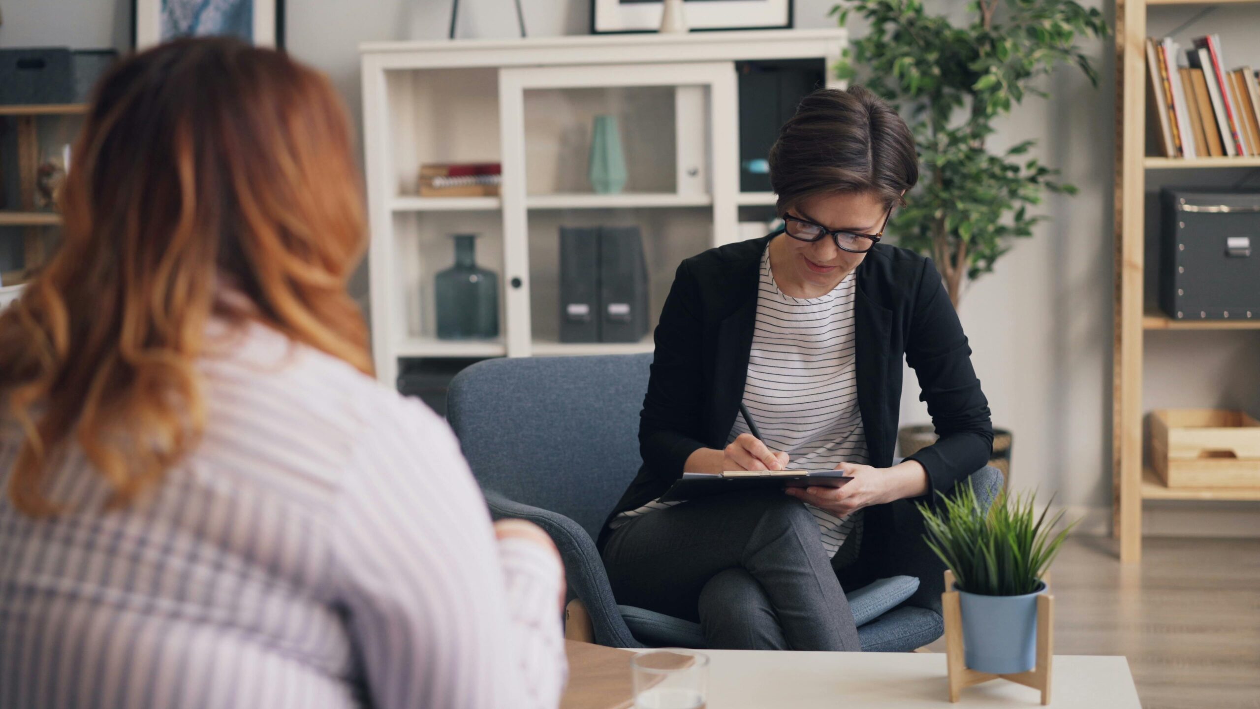 A female therapist wearing glasses and a dark blazer holding a clipboard and taking notes during a session.
