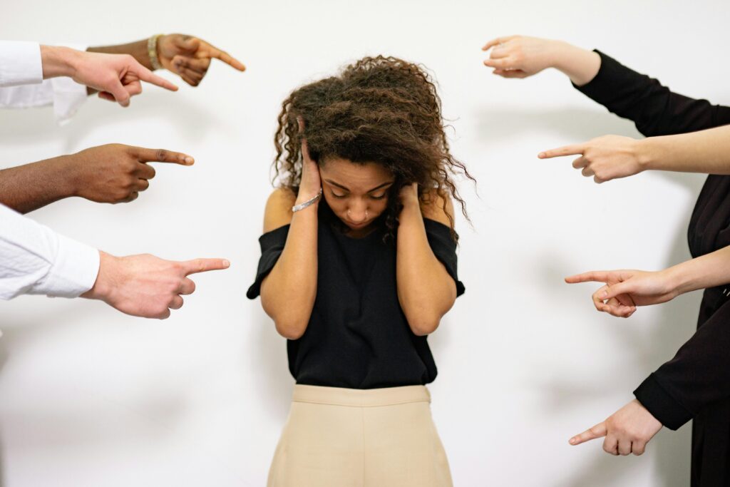 A woman in a black shirt covering her ears with her hands while several hands point toward her.