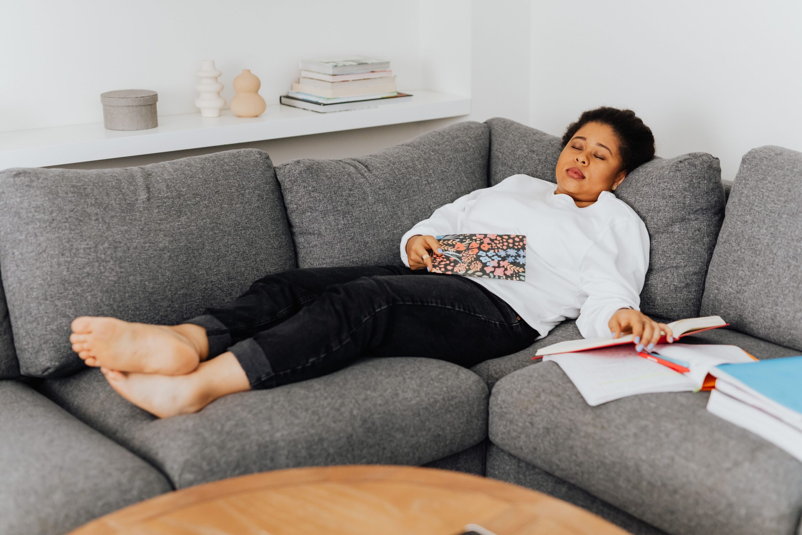 An exhausted, parentified woman resting on a grey sofa with open textbooks and papers nearby, holding a notepad as her nervous system finally downshifts from an anxious spiral with hypnosis.