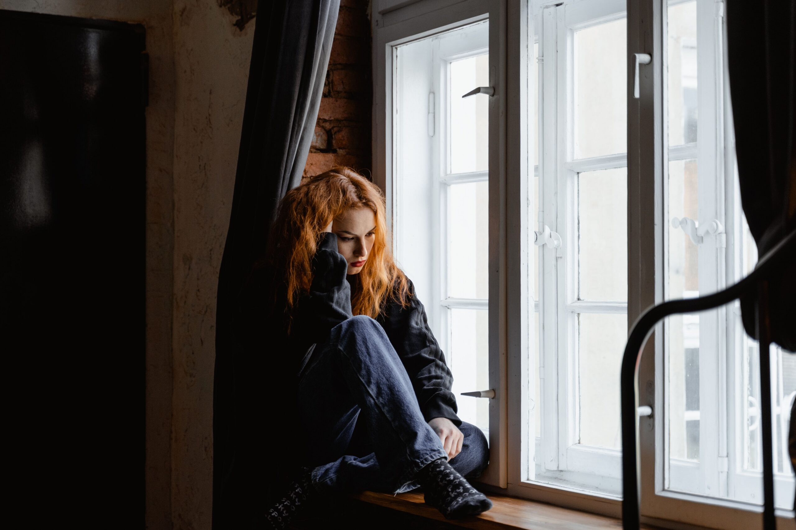 A young woman with auburn hair sits curled on a windowsill, looking down, in a dark moody room with natural light behind her, reflecting the emotional weight of wondering if therapy is working.